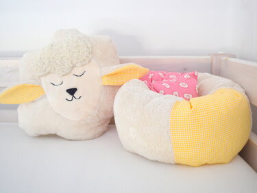 Plüsch-Lammkissen mit gestickten Augen neben rundem Babynest mit gelbem Karomuster und rosa Kissen auf heller Unterlage.