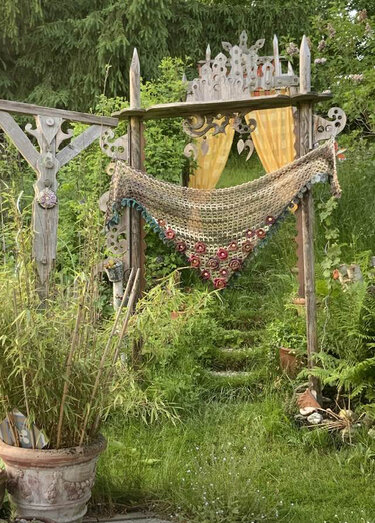 Beigefarbenes, handgehäkeltes Dreieckstuch mit roter Blumenborte an einem geschnitzten Holzbogen im Garten