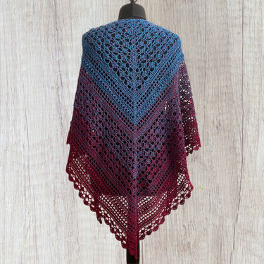 Crocheted triangular shawl with a blue-to-burgundy ombre, draped on a dress form against a wooden backdrop.