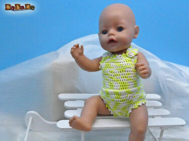 Babypuppe sitzt auf einem kleinen weißen Schlitten und trägt einen gehäkelten Strampler in Limettengrün und Weiß.