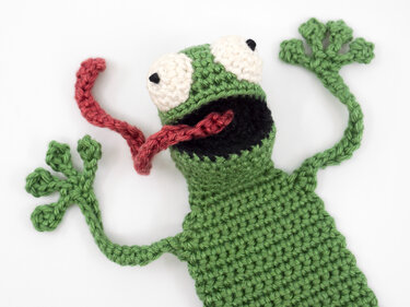 Green crocheted frog bookmark with white eyes and red tongue on white background