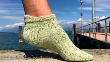 Handgestrickte hellgrüne Knöchelsocke mit Ajourmuster an einem Fuß vor Steg und See