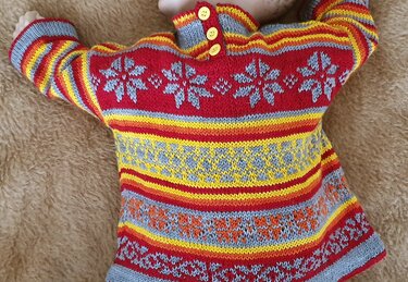 Gestrickter Baby-Pullover in Rot, Gelb, Orange und Hellgrau mit Schneeflocken- und Streifenmustern und drei gelben Knöpfen, auf beiger Decke liegend.