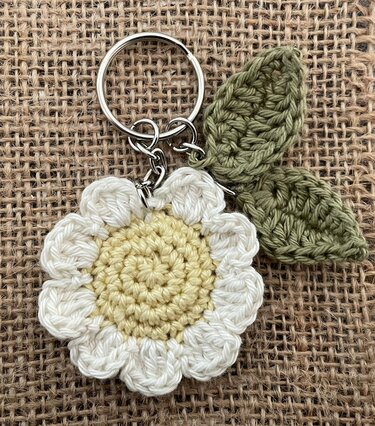 Nahaufnahme eines gehäkelten Gänseblümchen-Schlüsselanhängers mit cremeweißen Blütenblättern, hellgelber Mitte und zwei grünen Blättern an einem Metallring auf Jute.
