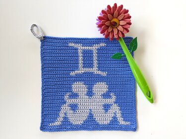 Blau-weißer gehäkelter Topflappen mit Zwillingssymbol und Aufhängeschlaufe, daneben grüner Küchenhelfer mit Blumenmotiv