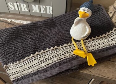 Dunkelgraues Frotteehandtuch mit cremefarbener gehäkelter Spitzenbordüre und dekorativer Möwenfigur mit blauer Mütze.
