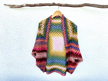 Bunter gehäkelter Seelenwärmer mit Granny‑Muster an einem Holzbügel vor heller Wand