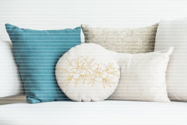 Rundes cremefarbenes Kissen mit goldener Blumen-Kontur zwischen türkis- und beigefarbenen Kissen auf einer Couch