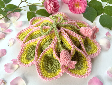 Gehäkelter Blütenbeutel in Pastellrosa, Hellgelb und Salbeigrün, dekoriert mit Rosenblüten und Blättern auf weißem Grund