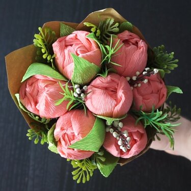 Hand hält einen Strauß aus rosafarbenen Krepppapierblüten mit grünen Blättern und braunem Papierumschlag vor dunklem Hintergrund.