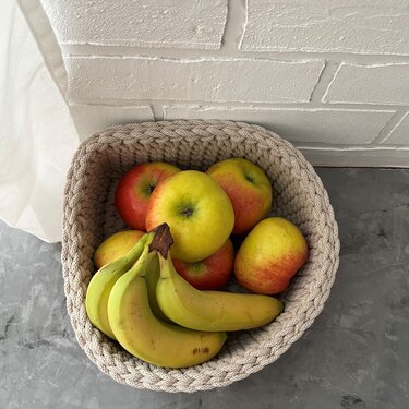 Beiger gehäkelter Korb, gefüllt mit Äpfeln und Bananen, auf grauer Fläche vor weißer Ziegelwand