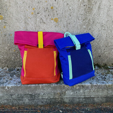 Zwei Rolltop-Rucksäcke in Pink-Orange und Blau-Dunkelblau auf einer Betonmauer