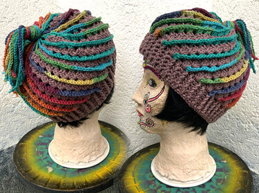 Gehäkelte Mütze mit braunem Grund und bunten Streifen sowie seitlichen Zöpfen auf zwei Schaufensterpuppen