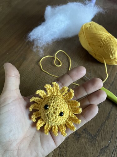 Hand hält kleine gelbe gehäkelte Sonne mit Augen; im Hintergrund Garn und Füllwatte