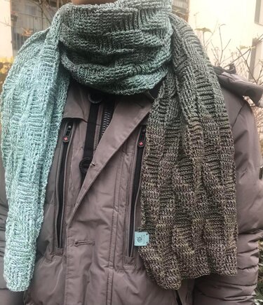 Person trägt einen gehäkelten Schal mit Farbverlauf in Grün- und dunklen Grüntönen und Reliefstruktur über einer grauen Jacke.