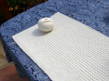 White knitted blanket or sack in progress with a ball of white yarn on a blue paisley tablecloth.