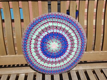 runder gehäkelter Kissenbezug mit konzentrischen Ringen in Blau, Mint, Rosa und Grau auf Holzbank