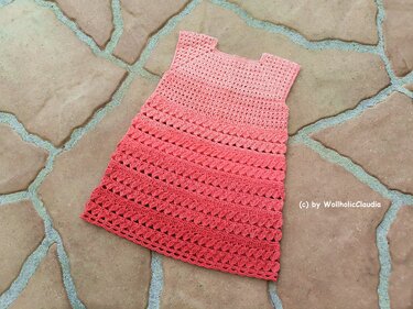 Ärmelloses gehäkeltes Babykleid in Rosa bis Korall, flach auf Steinboden gelegt, mit Loch- und Muschelmuster