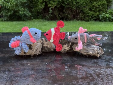 Drei gehäkelte Amigurumi-Fische (blau, rot, grau) mit pinken Akzenten auf Steinen arrangiert.