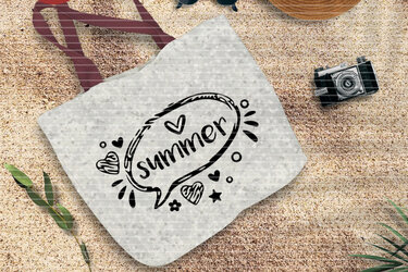 Helle bedruckte Stofftasche mit dem Schriftzug 'summer' liegt auf sandigem Boden, umgeben von sommerlichen Gegenständen.