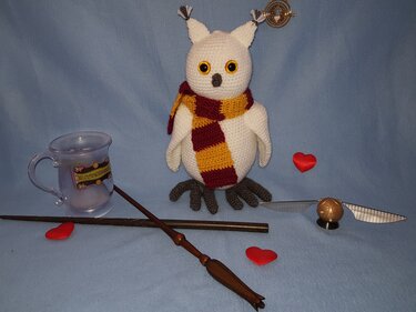 Weißes gehäkeltes Eulen‑Amigurumi mit gelb‑rotem gestreiftem Schal, umgeben von Zauberstäben, einer goldfarbenen Kugel mit Flügeln, einer transparenten Tasse und roten Herzen auf blauem Hintergrund.