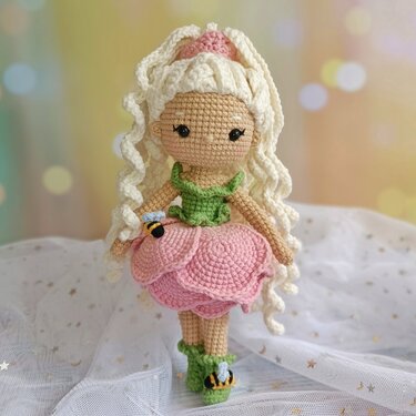 Amigurumi-Häkelpuppe mit cremefarbenen Locken, rosa Blütenrock, grünem Oberteil und Bienen-Applikationen