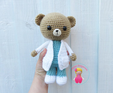 Brown crocheted amigurumi teddy bear wearing a white coat and teal scrubs, held by a hand.