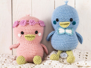 Zwei gehäkelte Amigurumi-Vögel, einer rosa mit Blumenkranz, der andere blau mit Fliege auf der Brust, sitzen auf einer Decke.