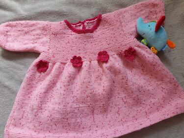 Rosa gestricktes Babykleid mit langen Ärmeln, gehäkelten dunkleren Blüten an der Taille, auf hellgrauem Untergrund liegend neben einem blauen Stoffelefanten