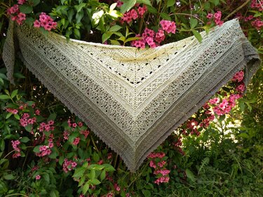 Gehäkeltes Dreieckstuch in hellen Beige- und Grüntönen mit Spitzendetails, aufgehängt an einem Strauch mit rosa Blüten.