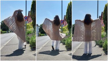 Person hält helle, gehäkelte Decke mit offenem Zickzack- und Rautenmuster auf einem sonnigen Gehweg.