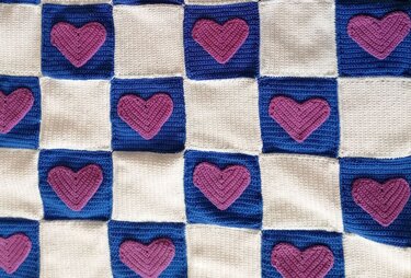 Gehäkelte Babydecke mit blau-weißem Schachbrettmuster und lila Herzapplikationen auf den blauen Feldern