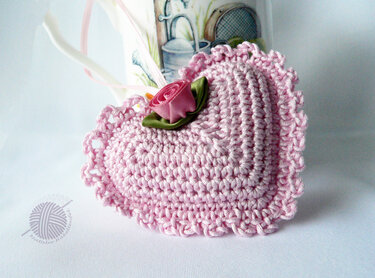 Helles rosa gehäkeltes Herz mit Rüschenrand und kleiner Satinschleifenrose auf hellem Untergrund.