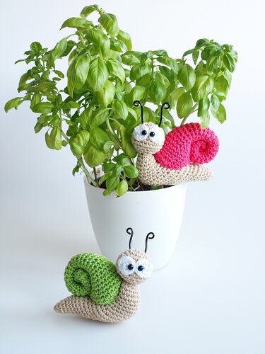 Zwei gehäkelte Amigurumi-Schnecken mit pinker und grüner Muschel neben einem Basilikumtopf
