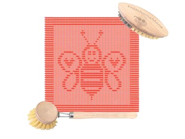 Rotes gestricktes Tuch mit großem Bienenmuster, daneben eine ovale und eine runde Holzbürste mit Holzgriff.