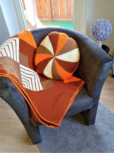 Rundes gestricktes Kissen in Orange, Braun und Creme auf dunkelgrauem Sessel mit gestrickter Blockstreifen-Decke