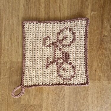 Quadratischer handgehäkelter Topflappen in Beige mit rosafarbenem Fahrradmotiv und Aufhängeschlaufe auf Holzuntergrund