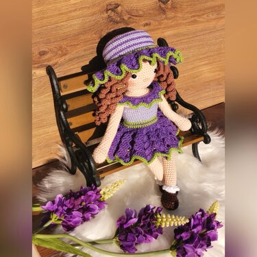 Crocheted amigurumi doll with purple ruffled dress and striped hat sitting on a wooden bench with purple flowers.
