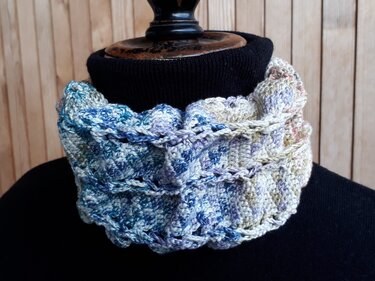 Gehäkelter Loop-Schal in Blau, Lila und Creme auf schwarzer Schaufensterpuppe vor Holzpaneelen.