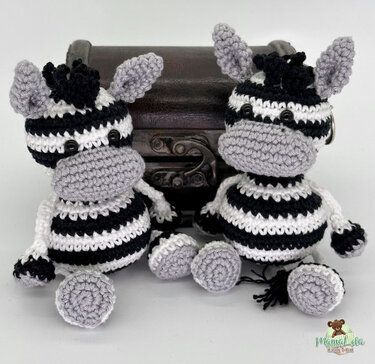 Zwei gehäkelte Zebra‑Amigurumi mit schwarzen und weißen Streifen, grauen Mäulern und Knopfaugen vor einer kleinen Truhe