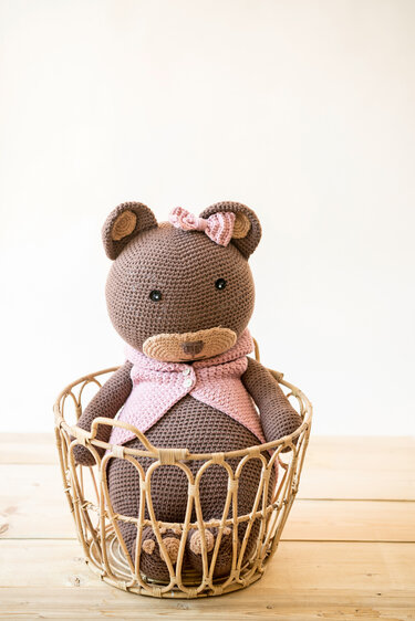 Brauner gehäkelter Teddybär mit rosa Weste und Schleife im Weidenkorb auf hellem Holz.