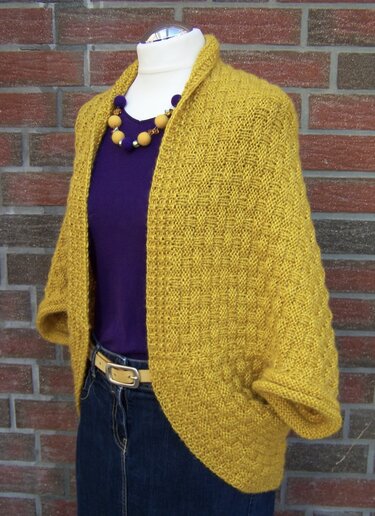 Mannequin trägt senfgelben, gestrickten Seelenwärmer über lila Top vor Ziegelwand