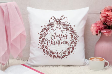 Weißes Kissen mit braunem Plott eines Weihnachtskranzes und dem Schriftzug 'Merry Christmas', daneben rosa Dekoration und eine Tasse Kaffee.