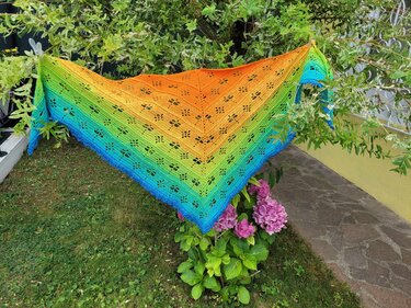 Buntes gehäkeltes Dreieckstuch mit Farbverlauf und libellenähnlichem Lochmuster, aufgehängt an Zweigen neben rosa Hortensien.