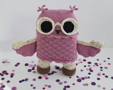 Gehäkelte Amigurumi-Eule in Lila und Creme vor hellem Hintergrund, umgeben von lila Knöpfen.