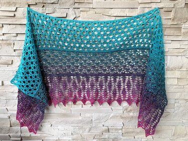 Crocheted shawl in teal, purple, and fuchsia ombre with a lacy openwork pattern and scalloped edge on a stone wall.