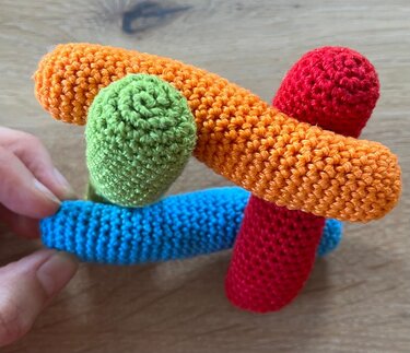 Vier gehäkelte Röhren in Rot, Orange, Blau und Grün; eine Hand hält das blaue Element
