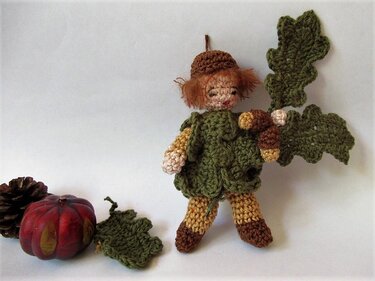 Gehäkelte Amigurumi-Waldwichtel-Puppe in grüner Kleidung mit Eicheln, Blättern, kleinem Kürbis und Kiefernzapfen.