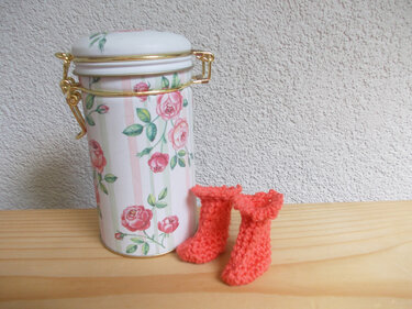 Zwei korallenfarbene gehäkelte Mini-Schühchen neben einer weiß-rosa Dose mit Rosenmotiv und goldfarbenen Verschlüssen.