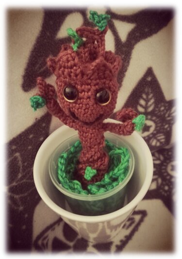 Gehäkelte braune Pflanzenfigur mit großen Augen und grünen Blättern in einem Topf, der in einer weißen Tasse steht.